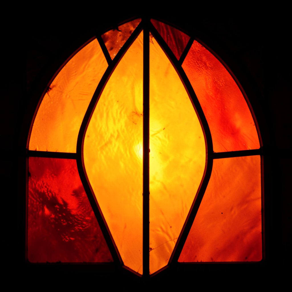 A wide horizontal banner in muted earth tones — sand, clay, and deep forest green — with a stained-glass-style segmented composition of bold leaded shapes evoking a Pacific island horizon and warm sunset light.